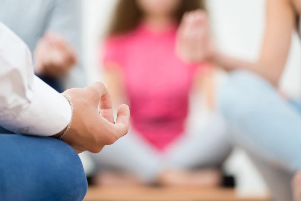 Personer som mediterar tillsammans. Närbild på en person som sitter i lotusställning. Fokus är riktat mot personens hand som hålls i en mudra. I bakgrunden syns suddigt tre personer som sitter mittemot på samma sätt. 